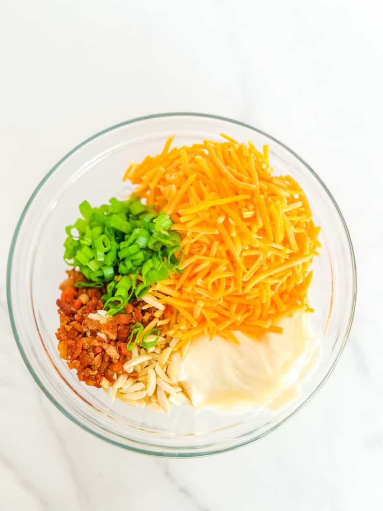 overhead photo of ingredients for Neiman Marcus Dip, including shreeded cheese, mayo, bacon pieces, slivered almonds and green onion in a clear mixing bowl