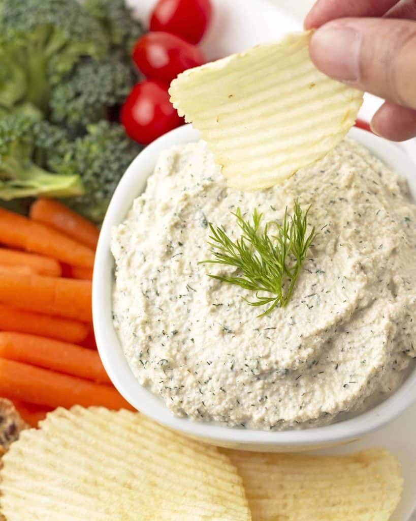 potato chip being dipped into vegan dip dip with veggies on the side