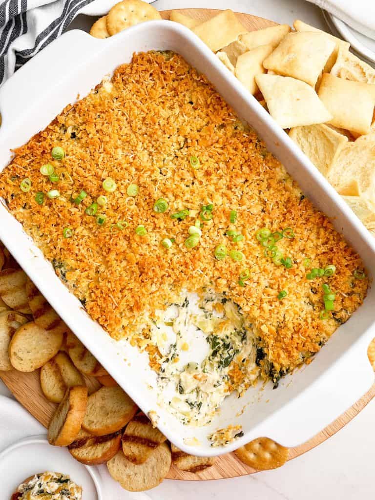 hot crab spinach and artichoke dip in a white casserole dish with dippers on the side