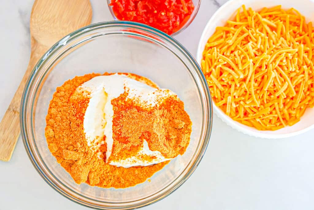 Overheat shot of bowlful of sour cream and taco seasoning next to bowls of shredded cheddar cheese and Rotel tomatoes