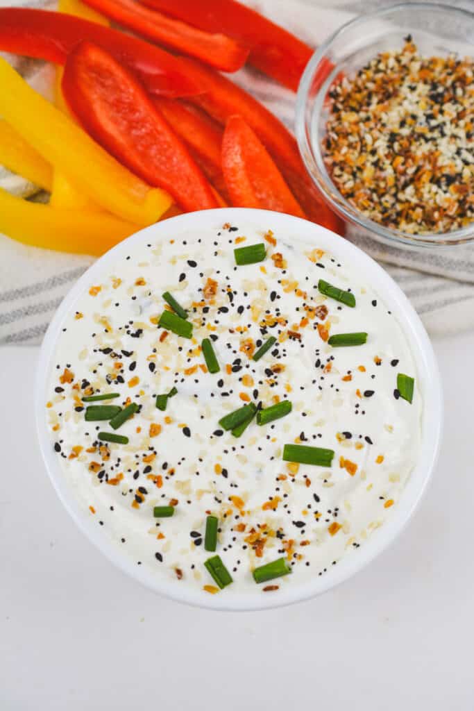 Overhead shot of bowlful of everything bagel dip next to bowl of more everything bagel seasoning and bell pepper strips