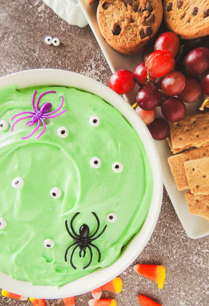 closeup of green halloween dessert dip with candy eyes surrounded by cookies, grapes, graham crackers and candy corn