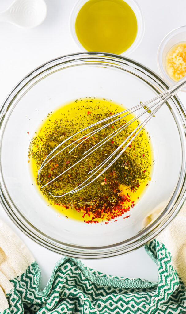 a whisk with bread dipping oil ingredients in a clear glass bowl