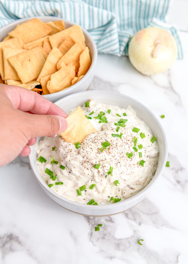 prepared caramelized onion dip in a white bowl with a pita chip being dipped by a caucasian man 