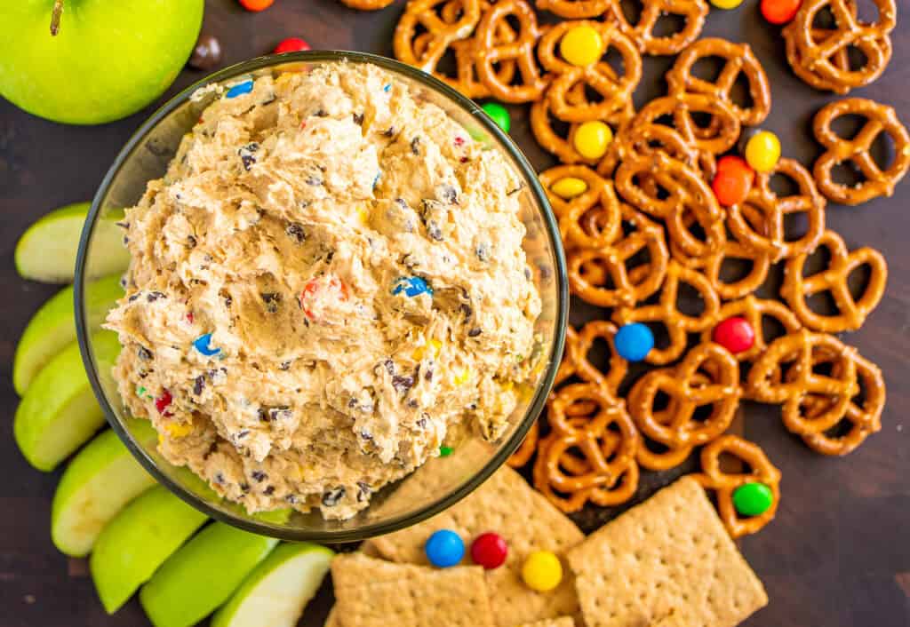 irresistible monster cookie dough dip in a bowl surrounded by m&m's candies, pretzels, apple slices, and graham crackers
