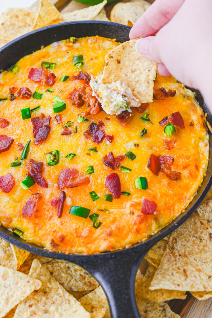 tortilla chip being dipped into prepared warm jalapeno popper dip in a cast iron skillet