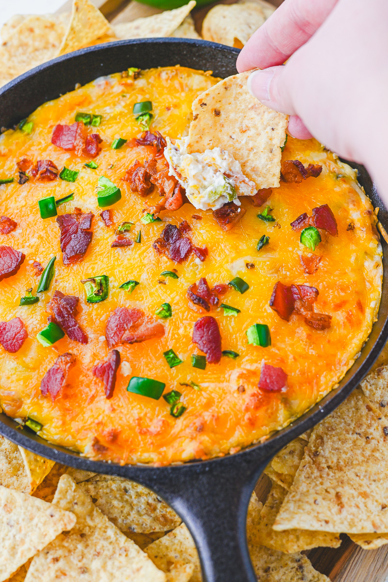 tortilla chip being dipped into prepared warm jalapeno popper dip in a cast iron skillet