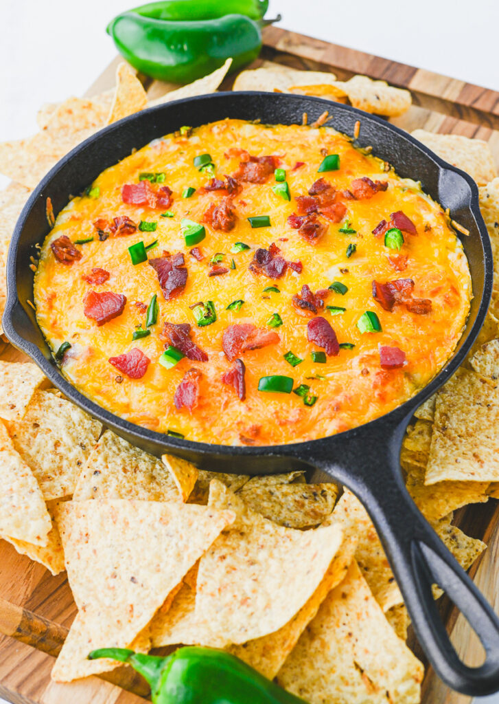 cast iron skillet with warm jalapeno popper dip garnished with bacon and jalapenos and a side of tortilla chips