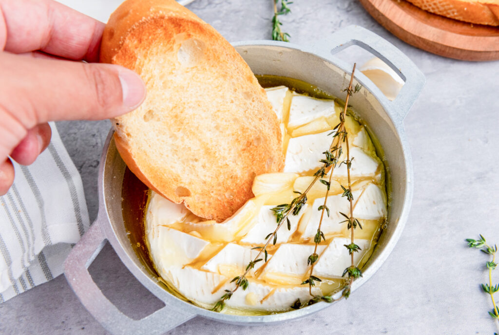 toasted baguette sliced being dipped into baked brie with honey and rosemary springs in a brie baker