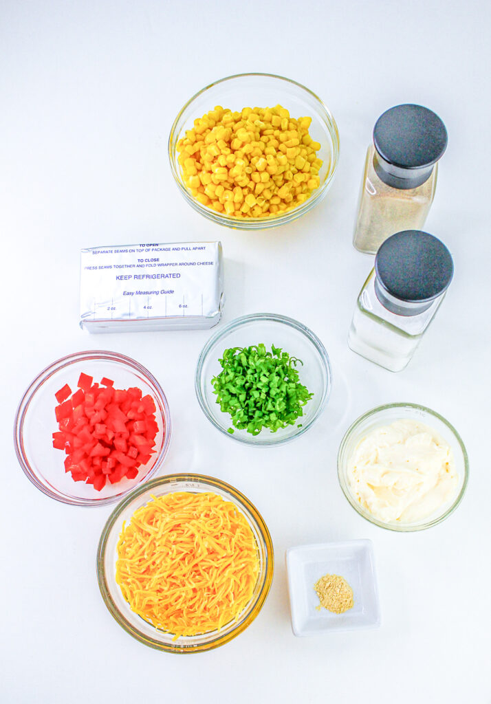ingredients laid out for hot corn dip, including cream cheese, kernaled corn, shredded cheese, red peppers, green onions, mayo, garlic and salt and pepper
