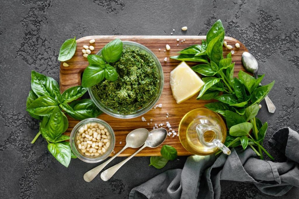 A jar of fresh green pesto with a mortar and pestle, basil leaves, and pine nuts nearby.