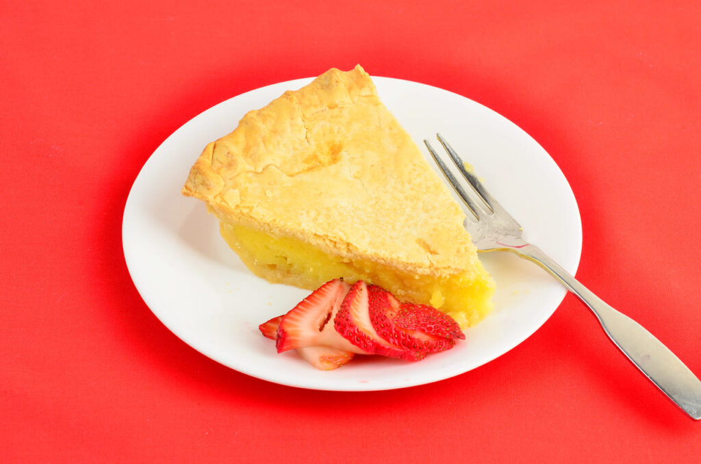 Slice of Shaker Lemon Pie with golden brown crust on white plate with sliced strawberry on red background.