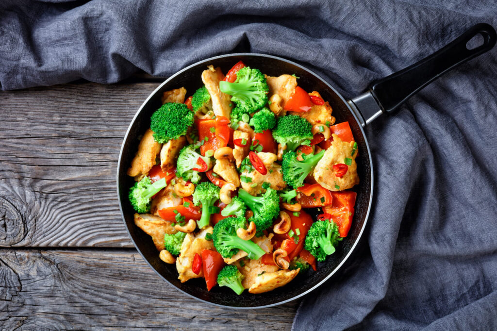 Cashew chicken with veggies stir-fry: broccoli, bell pepper, hot red chili, and soya sauce served on a skillet on a rustic wooden background, top view, close-up