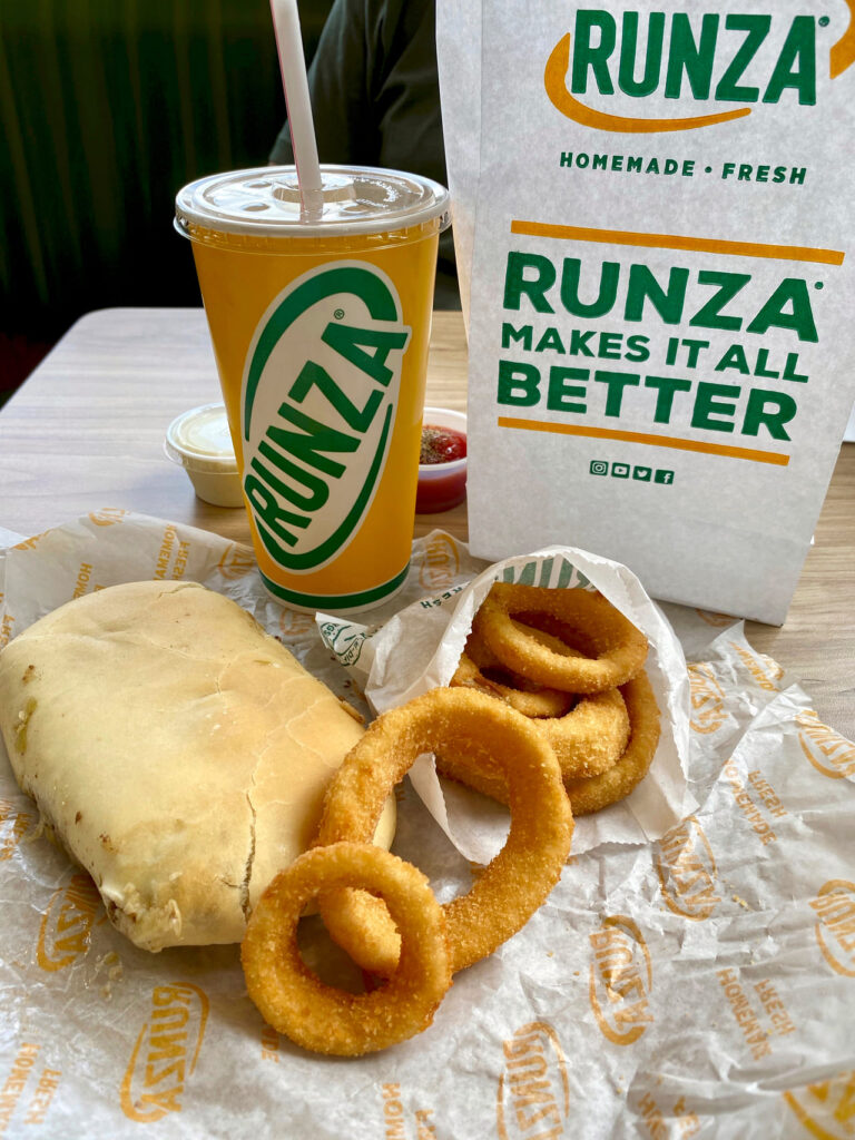 Runza Restaurant meal with a Runza sandwich, onion rings, and drink. Bag with Runza logo and "Runza makes it all better" motto. 