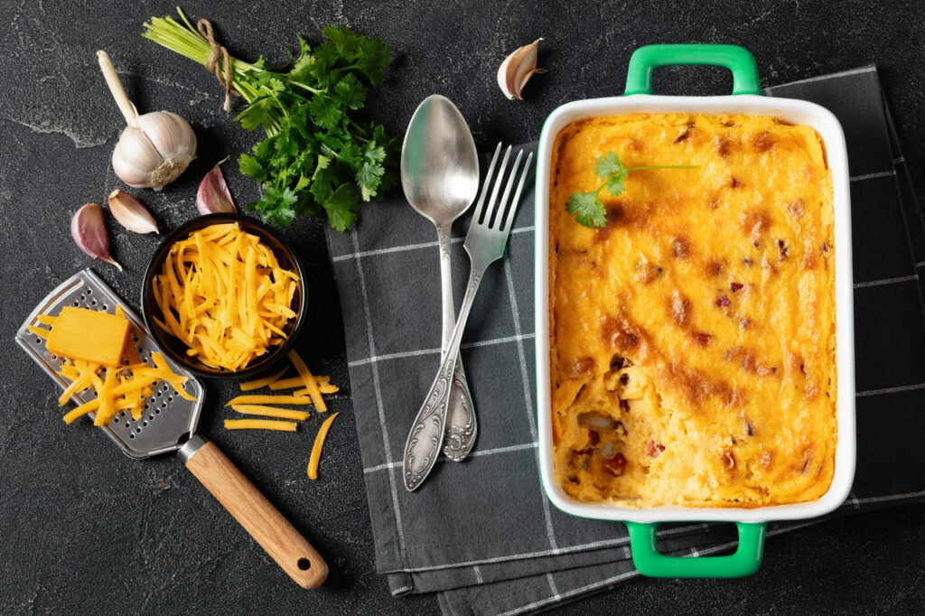 smoked sausage cheese grits casserole in green baking dish on dark concrete table with spoon and fork and ingredients, horizontal view from above, flat lay