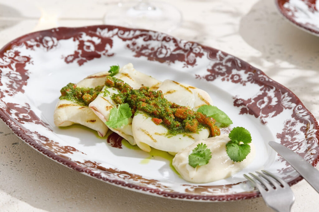Grilled squid with chimichurri sauce on vintage plate, bright natural lighting, angled composition.
