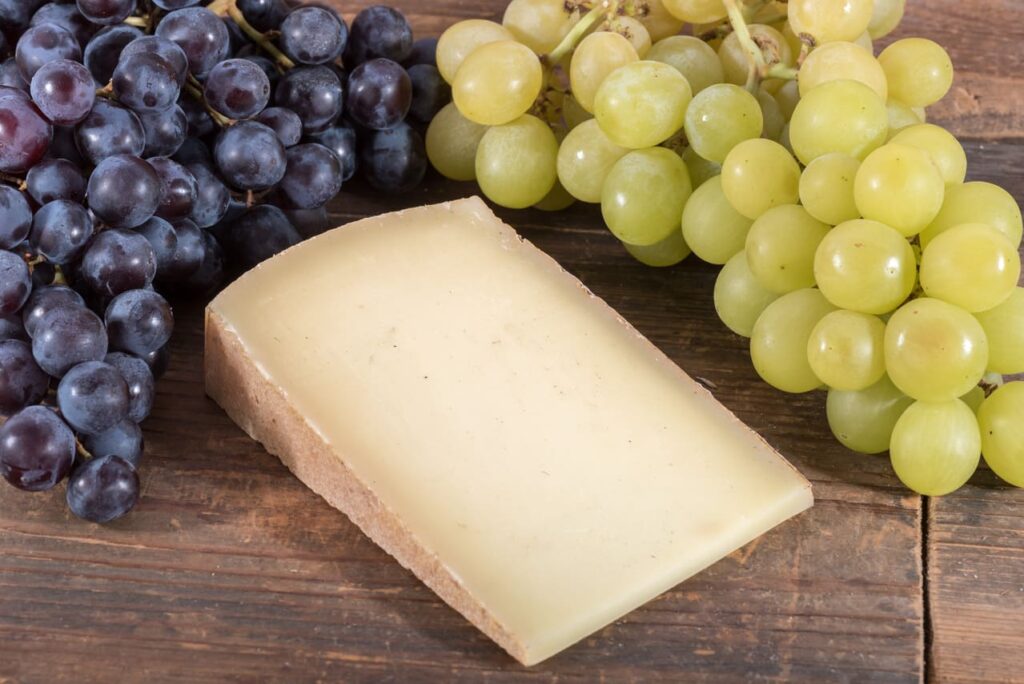French comté cheese with black and white grapes on wooden background
