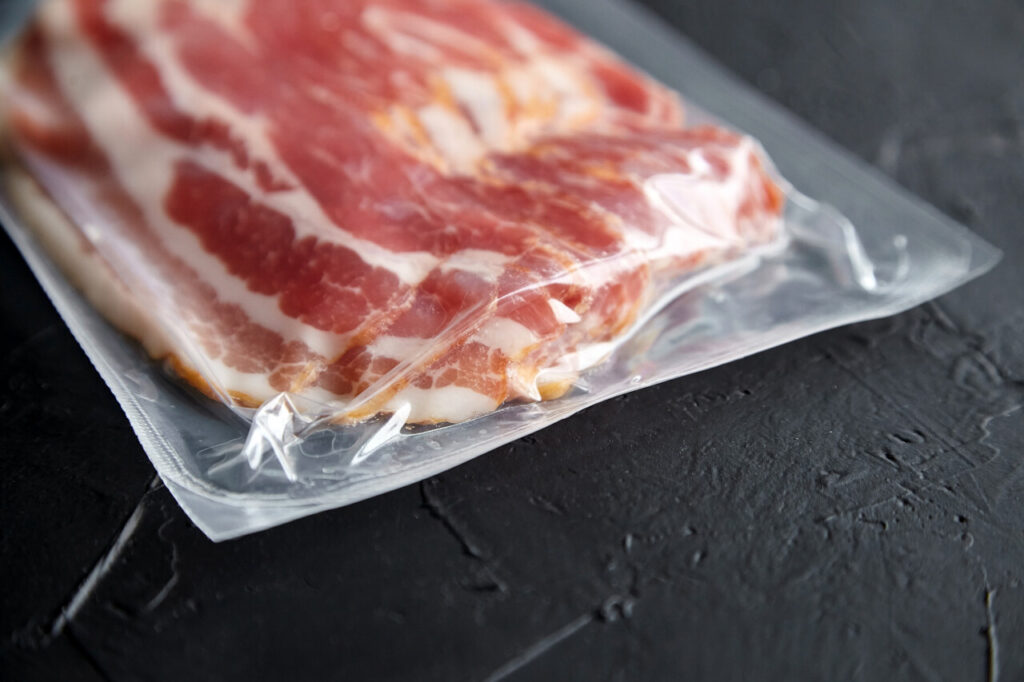 Bacon strips, raw smoked pork meat slices in vacuum package on black stone table