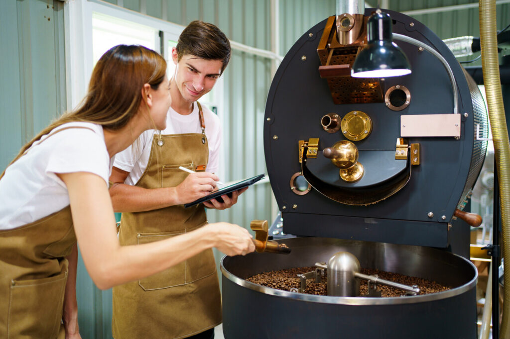 Male and female coffee roasters using the automatic coffee roasting machine roast an organic coffee beans. Coffee roasting specialist checking or inspecting quality of roasted coffee beans.
