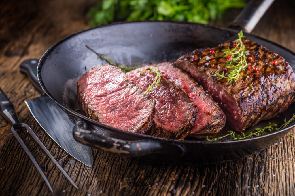 Beef steak. Juicy medium Rib Eye steak slices in pan on wooden board with fork and knife herbs spices and salt.