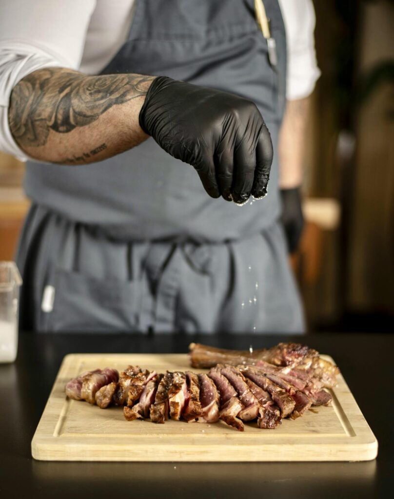 A chef sprinkling salt and pepper on raw steak.
