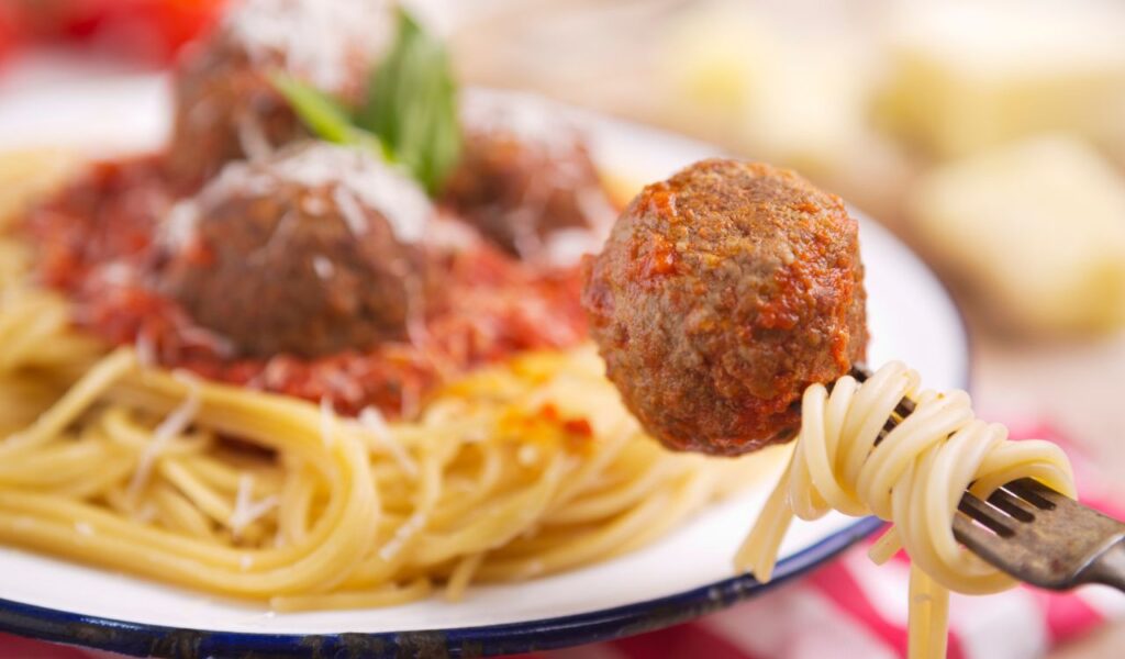 A plate with spaghetti with meatballs, topped with some grated parmesan cheese and basil. One meatball is picked up with a fork.