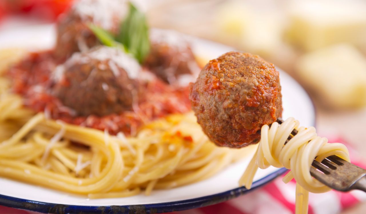 A plate with spaghetti with meatballs, topped with some grated parmesan cheese and basil. One meatball is picked up with a fork.